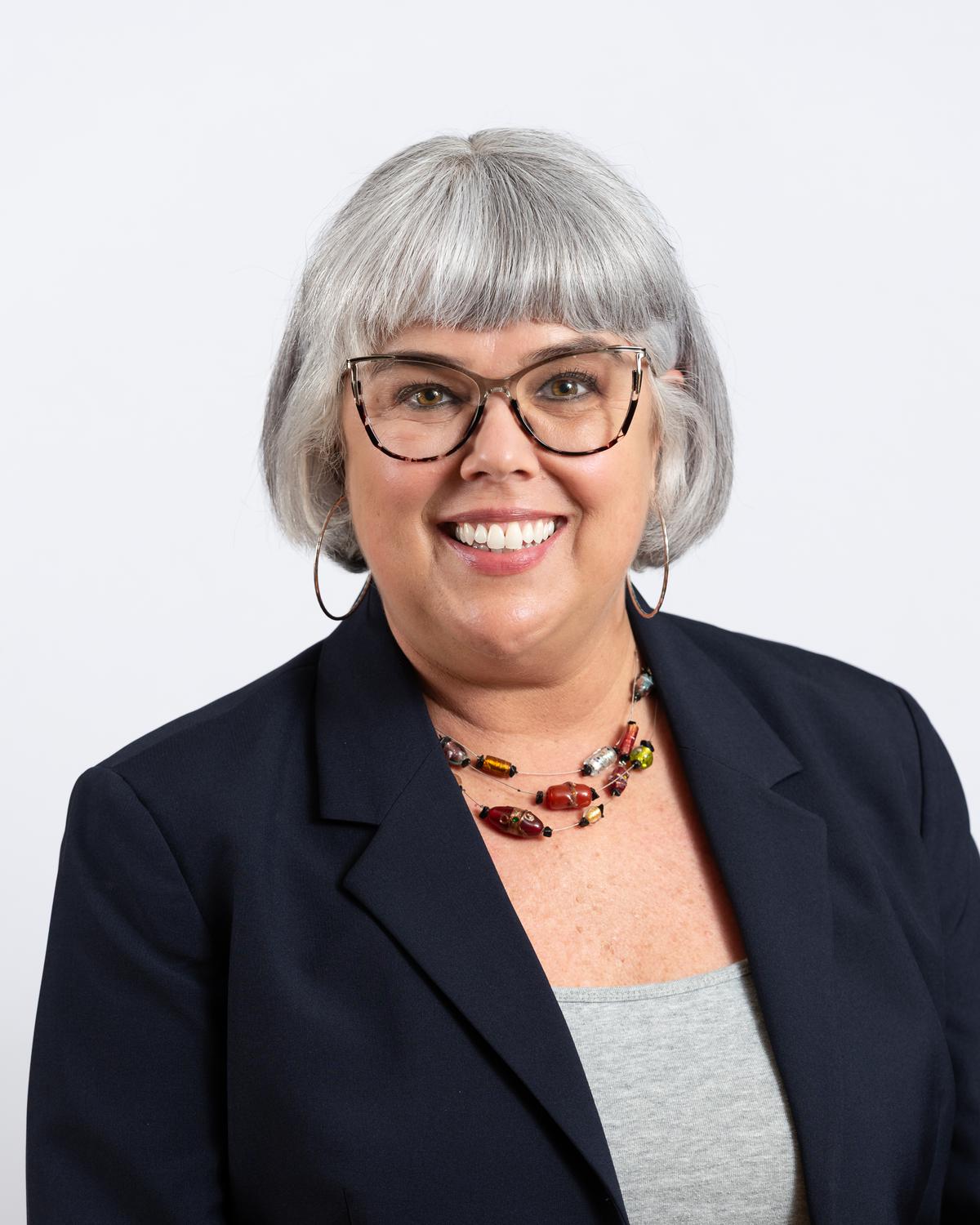 Corporate headshot of a woman in a black blazer and glasses against a white wall