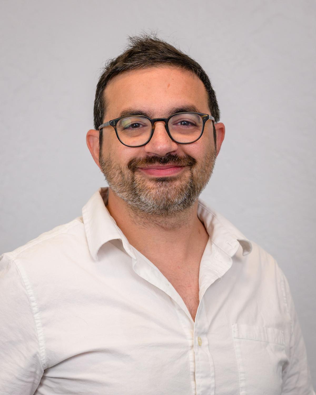 Headshot of a smiling man in glasses and a white open-collar shirt against a light grey backdrop