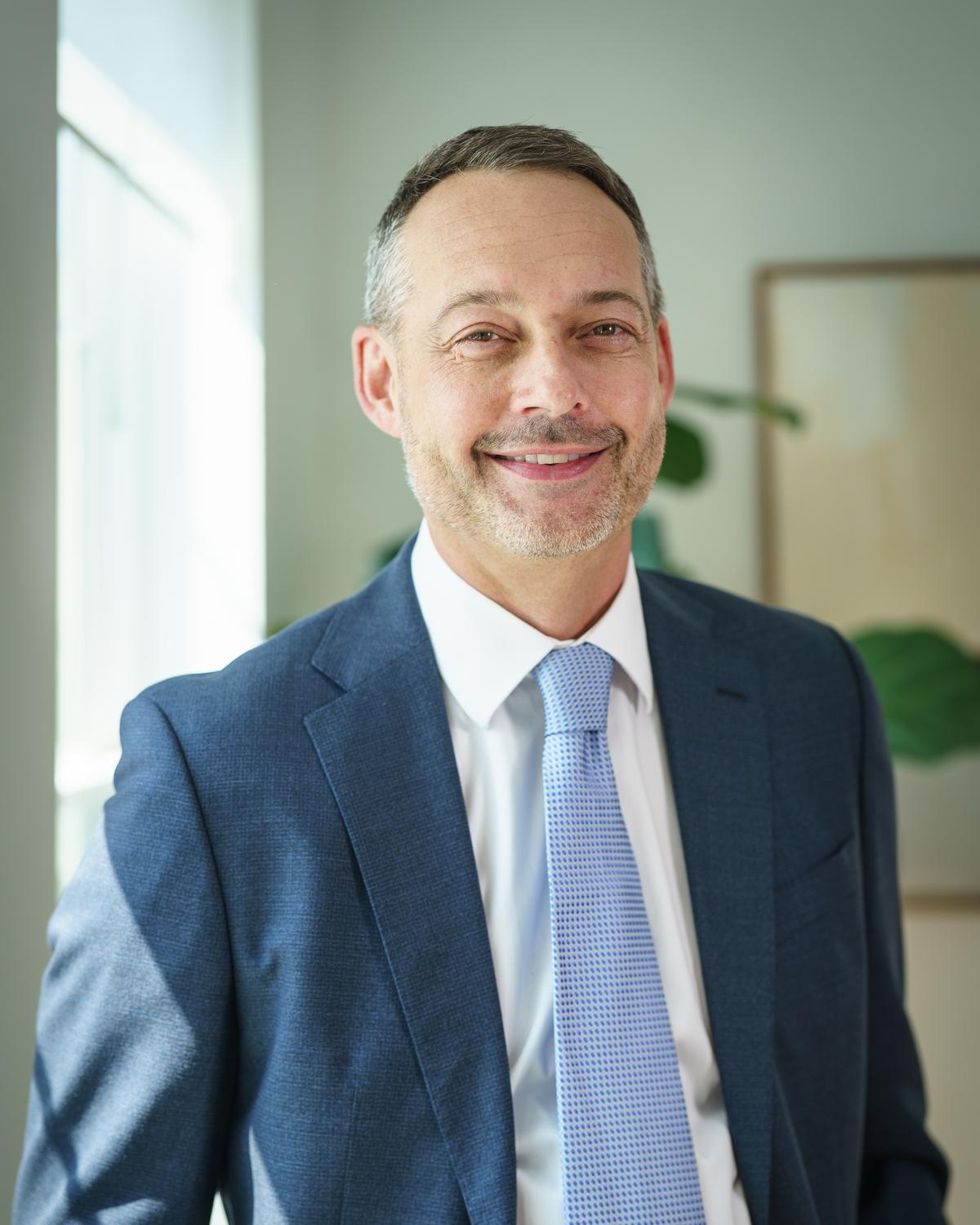 Corporate headshot of a man in a navy suit and blue tie in an office
