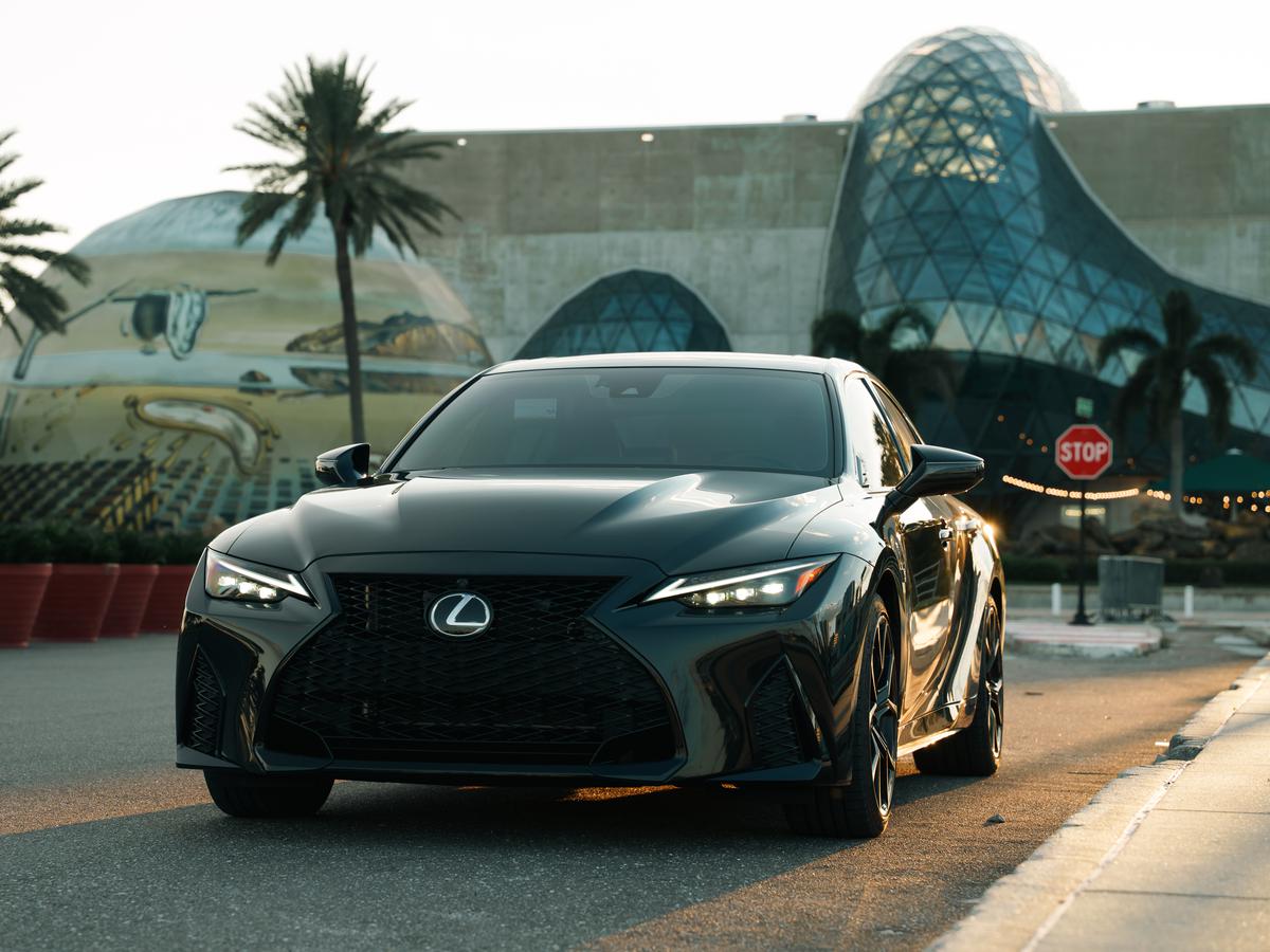 Dark green Lexus IS sedan with headlights on, parked on a palm-lined street at dusk