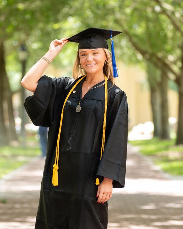 A graduate in cap and gown on a tree-lined path