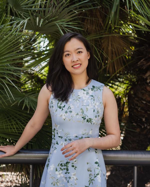 A smiling woman in a floral dress leaning on a railing among palm leaves