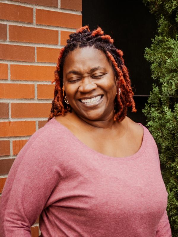 A smiling woman in a pink top beside a red brick wall