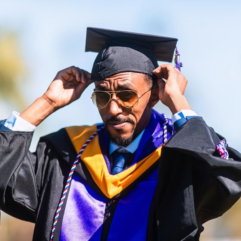 A graduate in gown and hood adjusting his cap against a blue sky