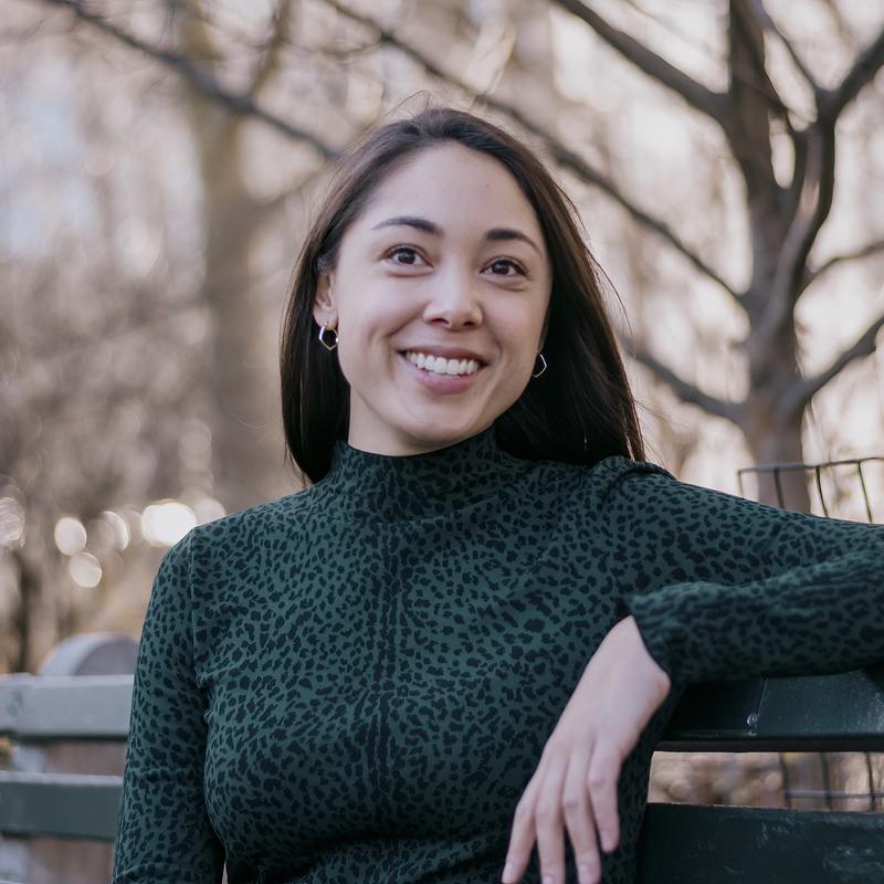 A smiling woman in a green sweater leaning on a bench outdoors