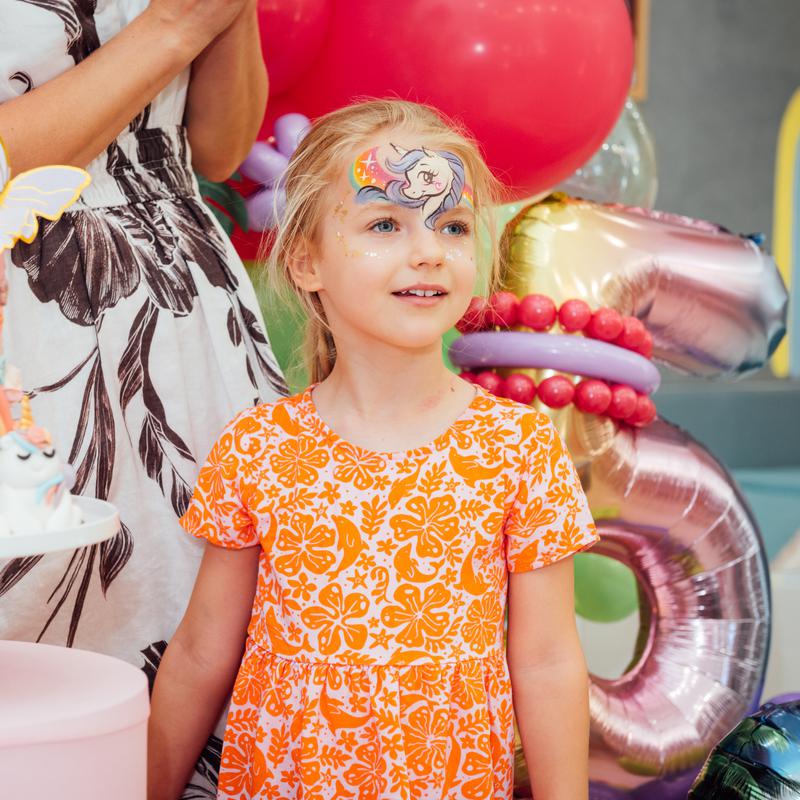 A young girl with face paint in an orange dress beside birthday balloons