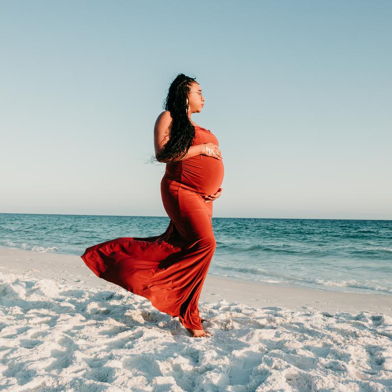An expectant mother in a long red dress on a white sand beach